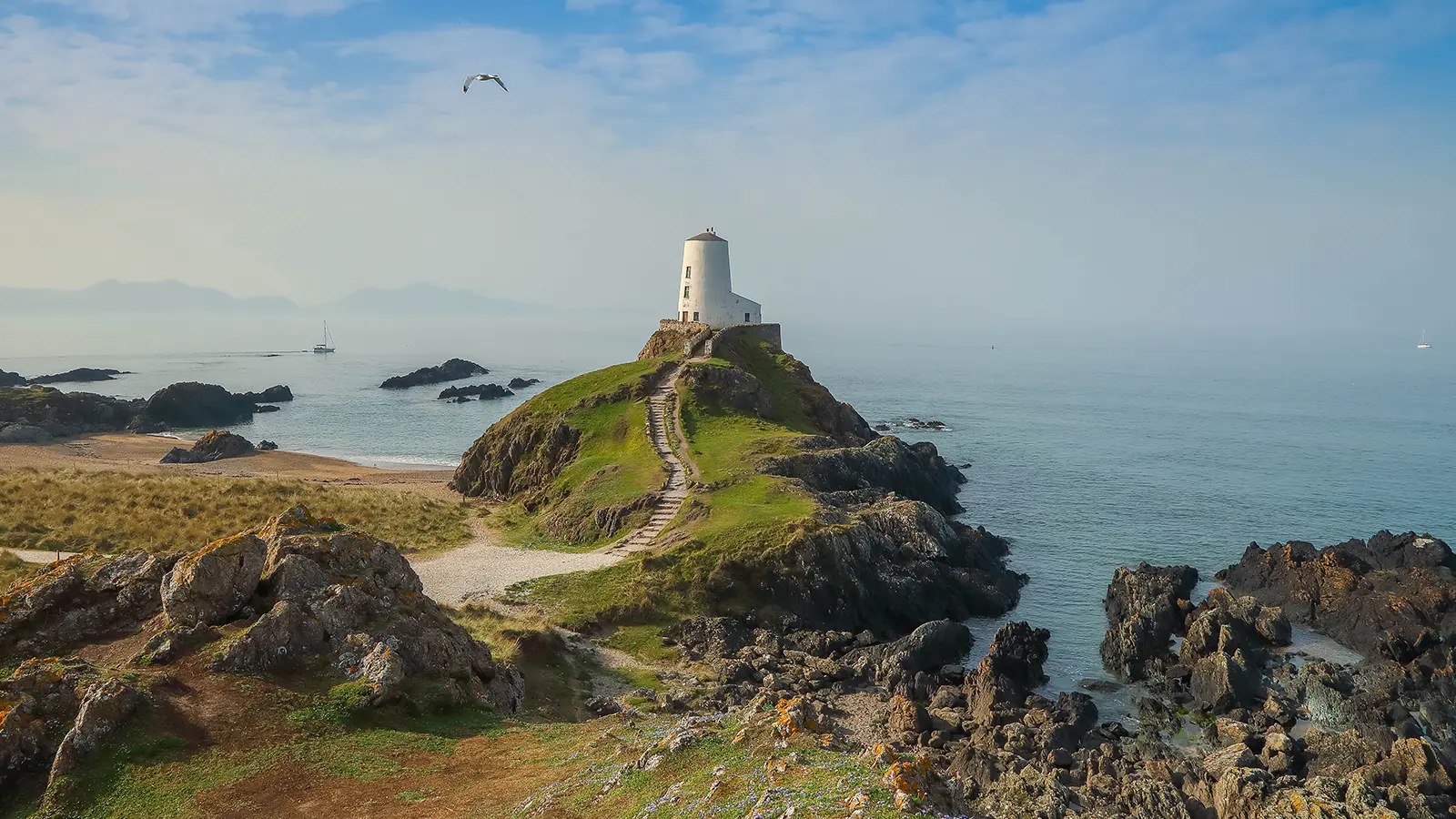 Coastal lighthouse on grassy cliff overlooking ocean with sailboats in distance