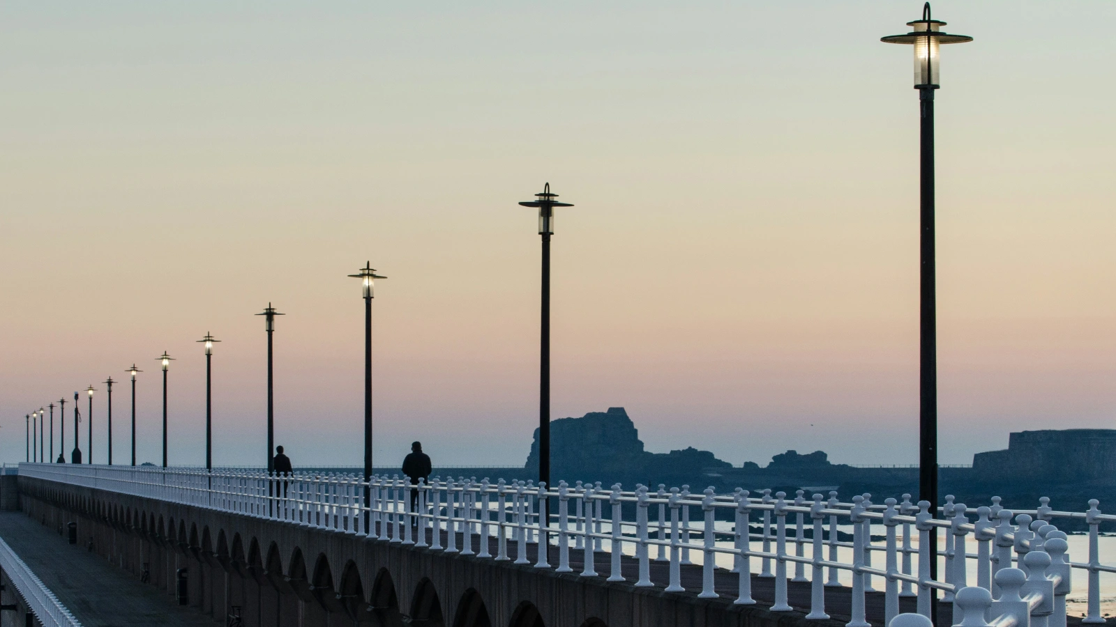 Jersey coastal promenade with evenly spaced lamps, two people walking beside calm water