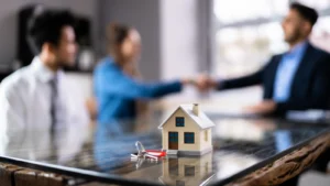 Small house model and keys on table with people shaking hands behind