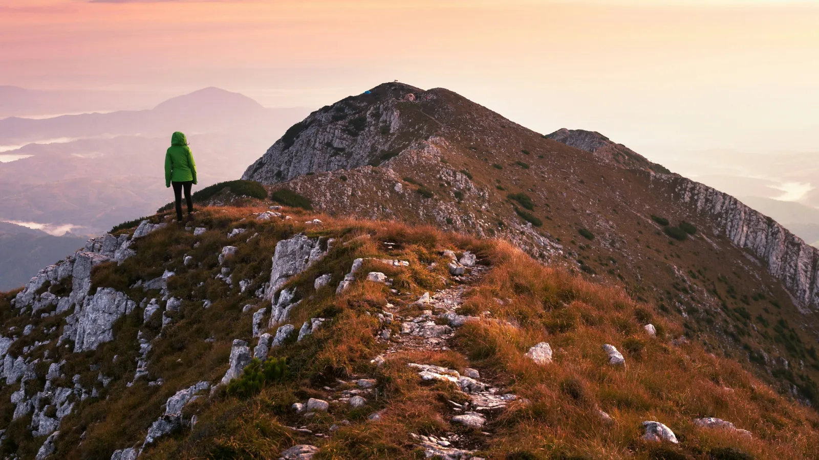 Person in green jacket standing on rocky ridge overlooking mountain range at sunset