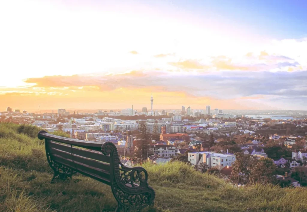 Wooden bench on a grassy hill overlooking a city skyline at sunset under a cloudy sky