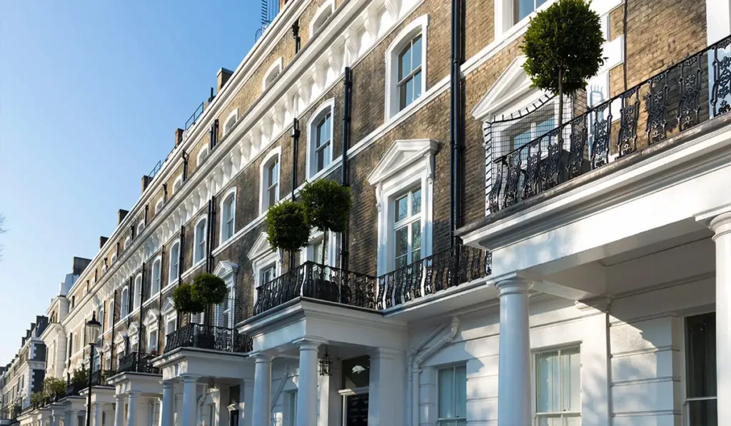 Row of white period London townhouses with black wrought-iron balconies and topiary trees under a clear blue sky