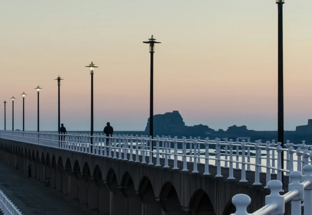 People walking along a balustraded seaside promenade lined with lampposts at dusk