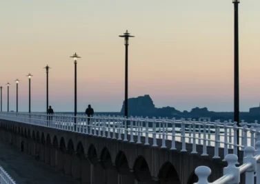 People walking along a balustraded seaside promenade lined with lampposts at dusk
